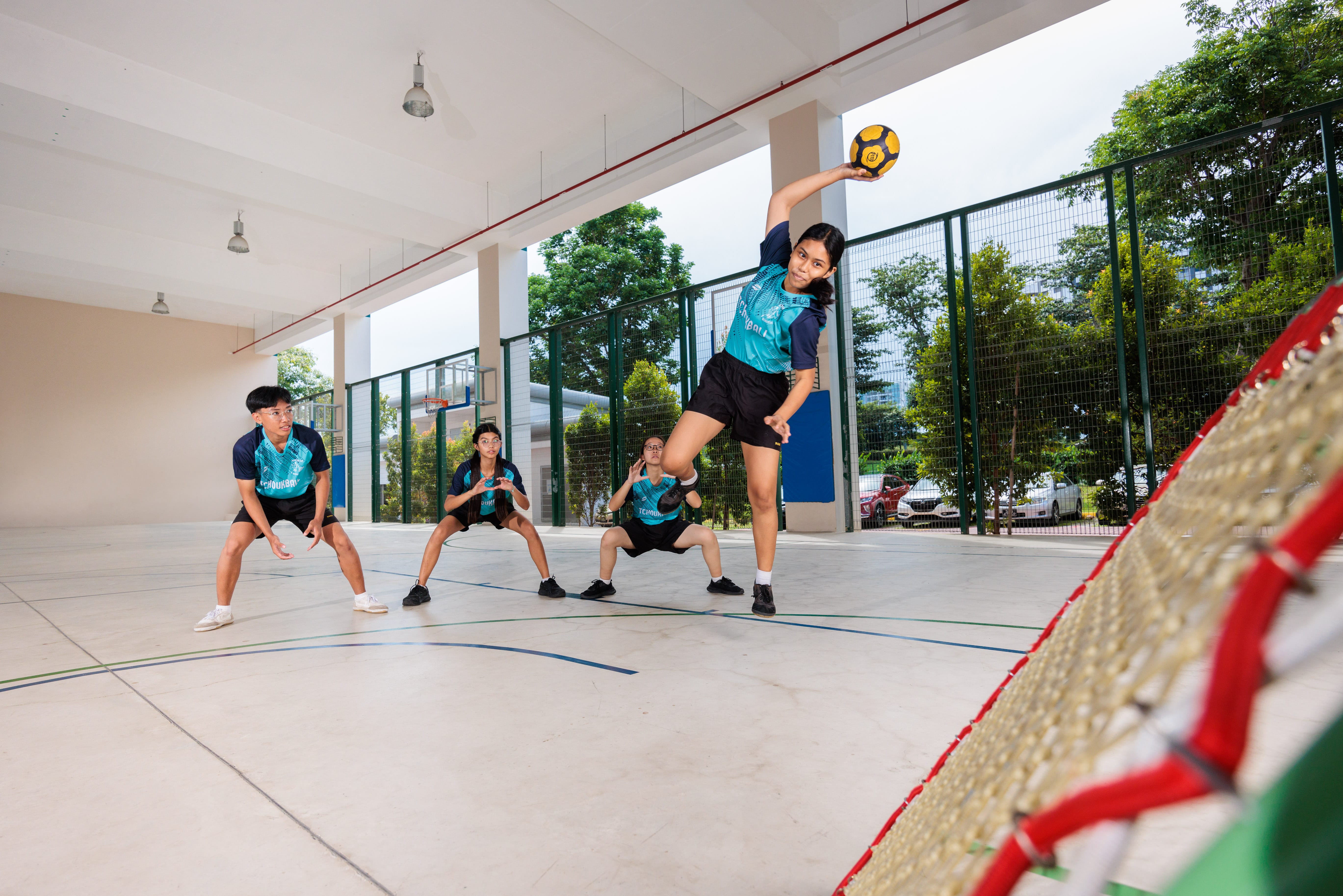 Four youths in teal jerseys play handball on an indoor court, one leaping to throw a yellow ball at a net.